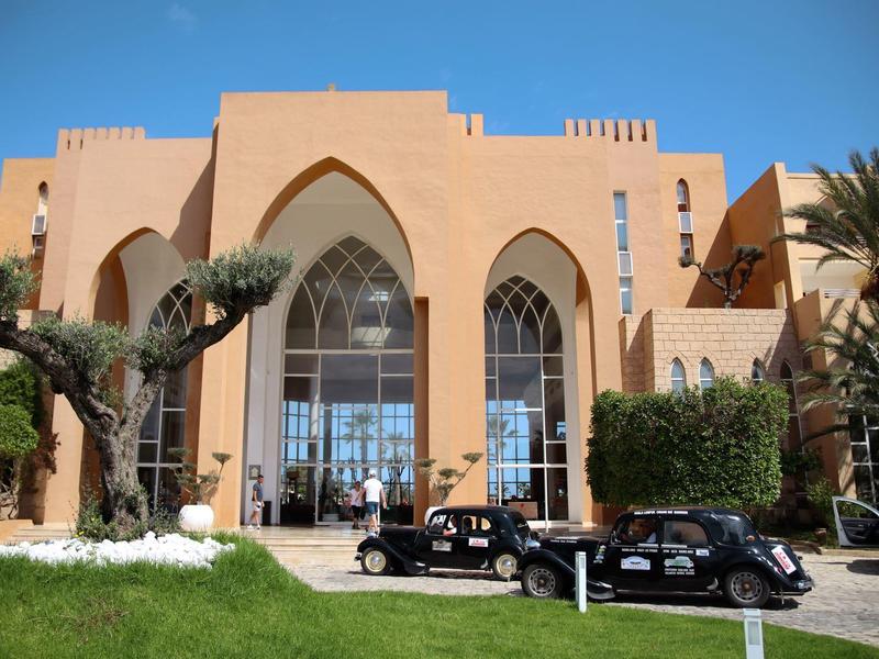 Velká hotelová budova v arabském architektonickém stylu, palmy a dvě veterány před ní.