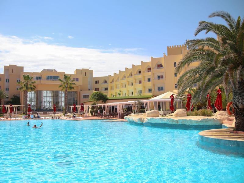 Velký venkovní bazén před béžovým hotelem s palmami a slunečníky.