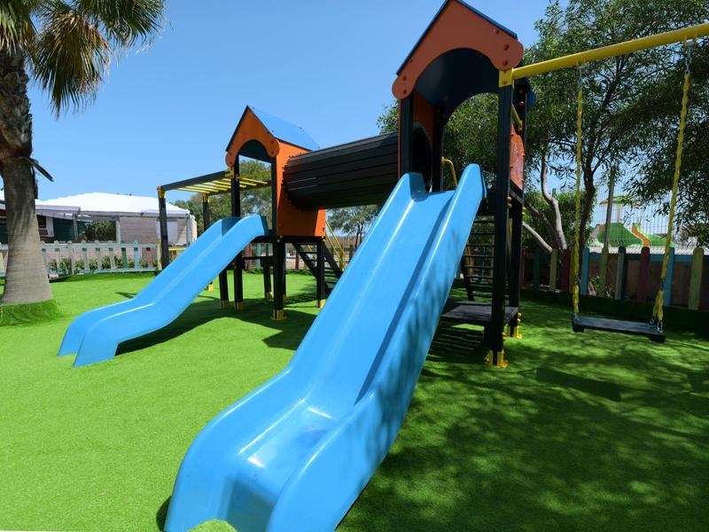 Dětské hřiště se skluzavkami a houpačkou na zelené umělé trávě pod modrou oblohou.