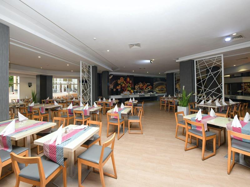 Světle osvětlená restaurace s dřevěnou podlahou, stoly s barevnými ubrousky a moderním dekorem.