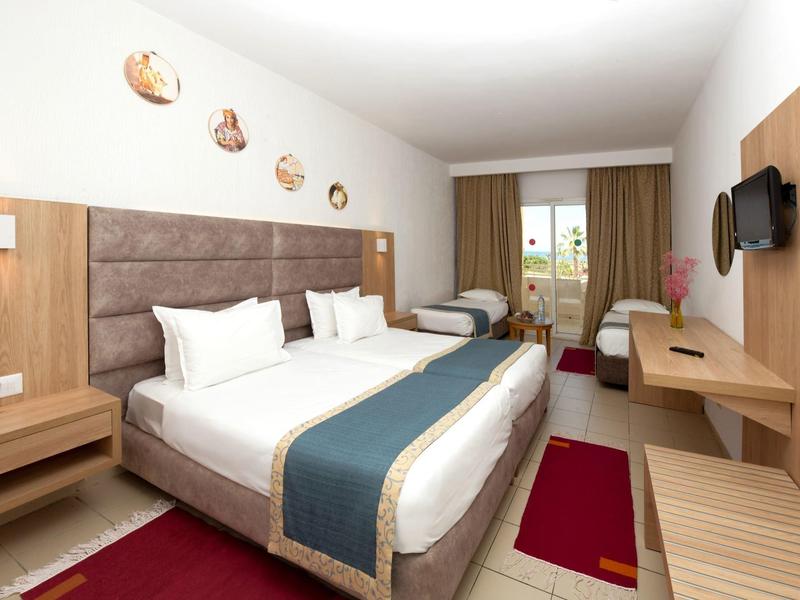 Hotelový pokoj s manželskou postelí, jednolůžkem, pracovním stolem a přístupem na balkón.