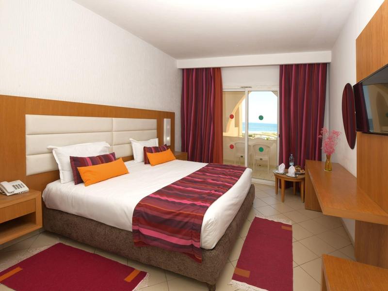 Moderní hotelový pokoj s manželskou postelí, balkonem, televizí a dřevěným nábytkem v teplých odstínech