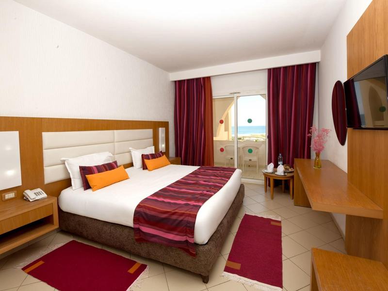 Moderní hotelový pokoj s velkou postelí, tmavě červenými závěsy a dřevěným nábytkem.