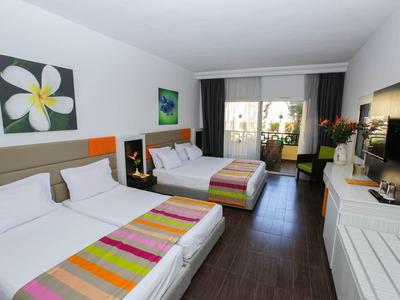 Hotelzimmer mit zwei Doppelbetten, bunten Tagesdecken, Fernseher und Balkon mit Pflanzen.