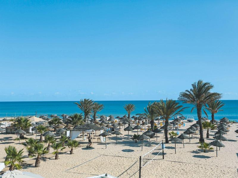 Strand mit Sand, Sonnenschirmen und Palmen am Meer unter blauem Himmel.
