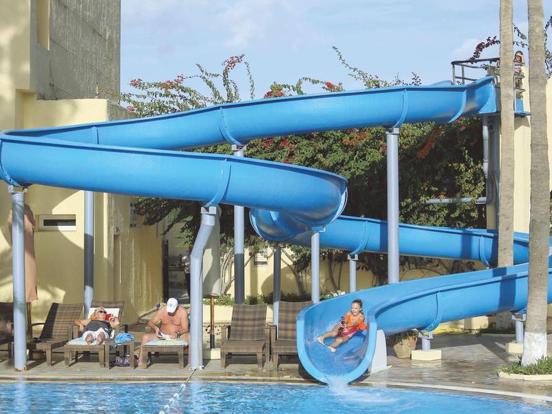 Kinder rutschen eine blaue Wasserrutsche in ein Schwimmbecken bei Sonnenschein.