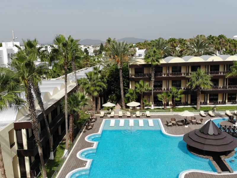 Großer Hotelpool mit Sonnenschirmen und Liegestühlen, umgeben von Palmen und Hotelgebäuden.