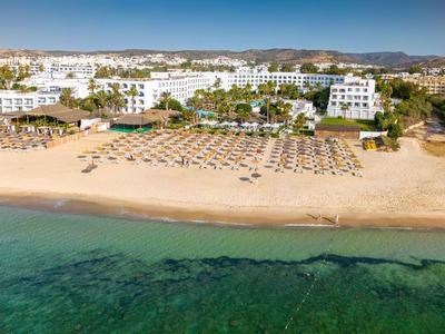 Strand mit Liegestühlen vor weißen Hotels und klarem türkisfarbenem Wasser unter blauem Himmel.