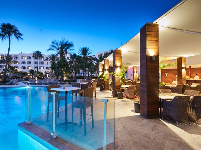 Moderner Außenbereich eines Hotels mit beleuchtetem Pool, Palmen und Sitzgelegenheiten bei Abenddämmerung.