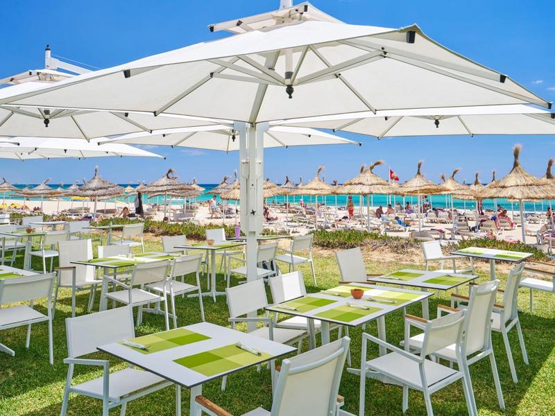 Strand mit Sonnenschirmen, weißen Tischen und Stühlen auf grünem Rasen bei klarem Himmel.