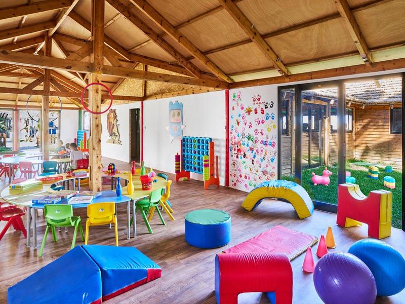 Bunter, heller Kinderraum mit Holzdach, kleinen Tischen, Stühlen und verschiedenen Spielgeräten.