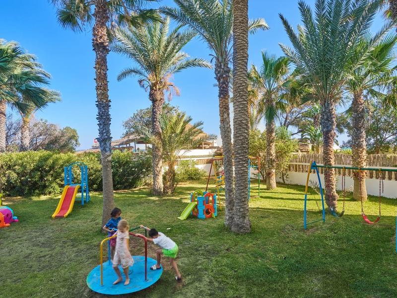 Kinderspielplatz mit Rutsche, Schaukel und Karussell, umgeben von hohen Palmen und grünem Gras.