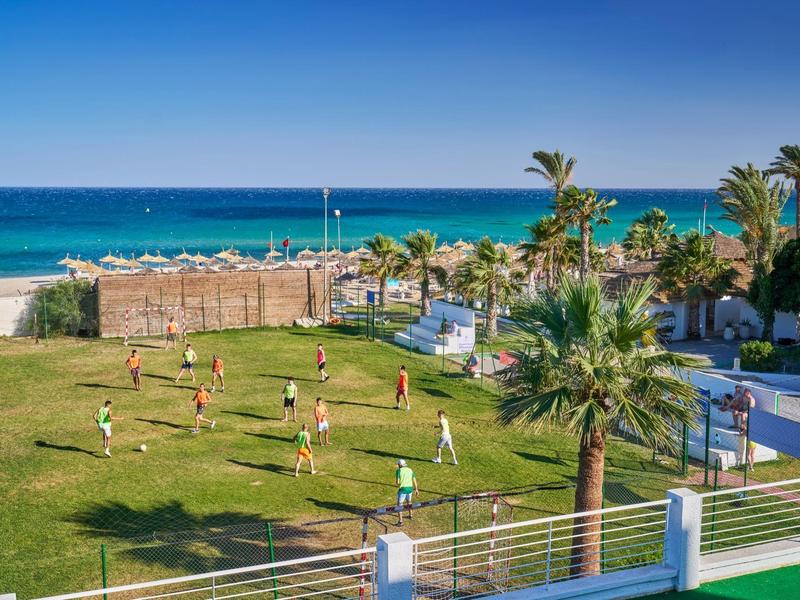 Strandnahes Spielfeld mit spielenden Kindern, Palmen und blauem Meer im Hintergrund.