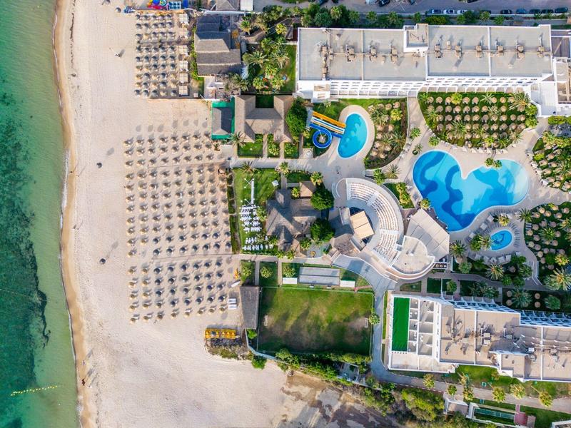 Luftaufnahme eines Resorts mit weißen Sandstrand, blauen Pools und grünen Gartenanlagen.