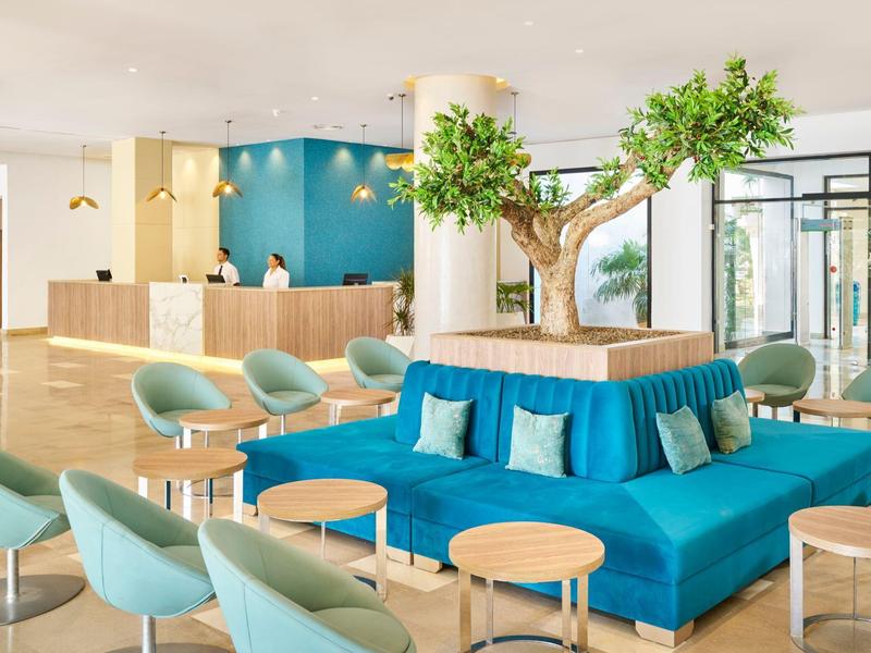 Moderner Empfangsbereich mit blauer Couch, grünen Sesseln, runden Tischen und großem Baum in der Mitte.