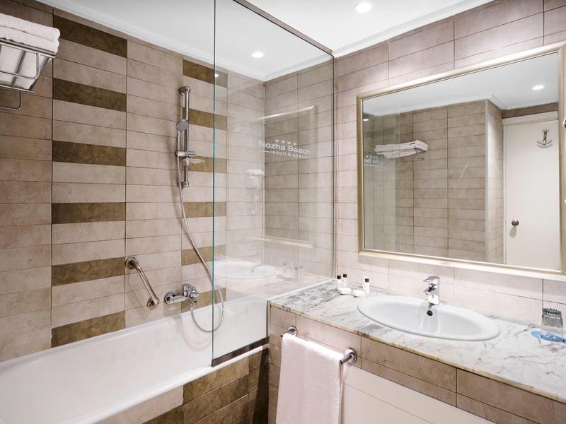 Helles Badezimmer mit Badewanne, Spiegel, Waschbecken und hellbraunen Fliesen an Wänden und Boden.