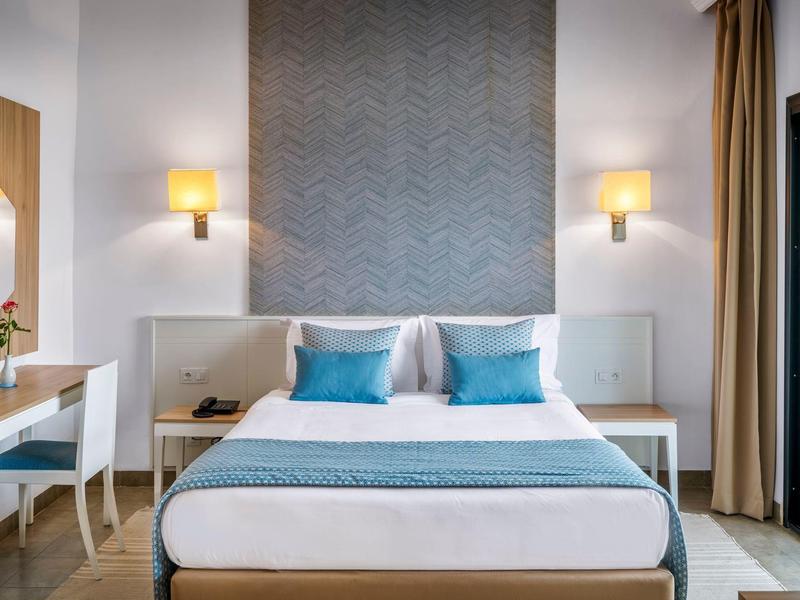 Modernes Hotelzimmer mit Doppelbett, blauen Kissen und Schreibtisch mit Stuhl.