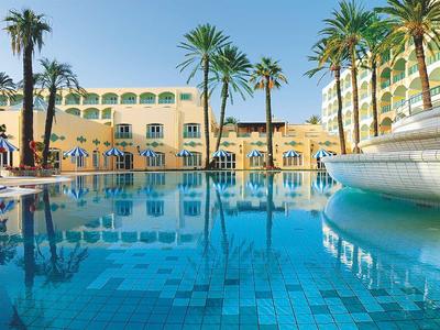 Großer blauer Pool vor gelben Hotels, gesäumt von hohen Palmen und Sonnenschirmen.