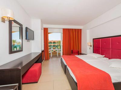 Modernes Hotelzimmer mit Doppelbett, rotem Bettkopfteil und Vorhängen, Schreibtisch und Balkon.