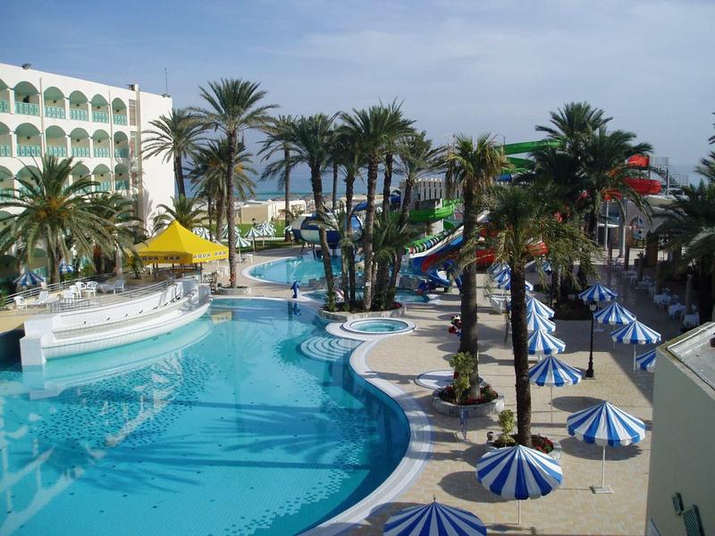 Großer Hotelpool mit Palmen, Wasserrutschen und blauen Sonnenschirmen an einem sonnigen Tag.