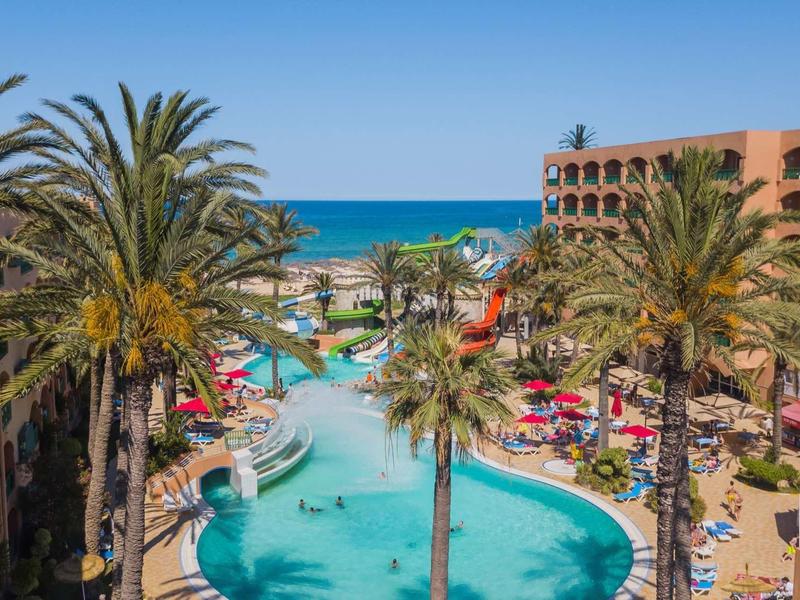 Großer Pool mit Palm Bäumen, roten Sonnenschirmen und Meer im Hintergrund bei klarem Himmel.