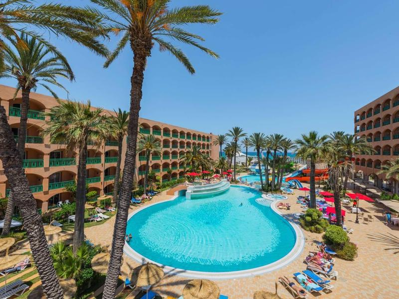 Großer, runder Pool mit Palmen und Liegen zwischen zwei mehrstöckigen Hotelgebäuden bei Sonnenschein.