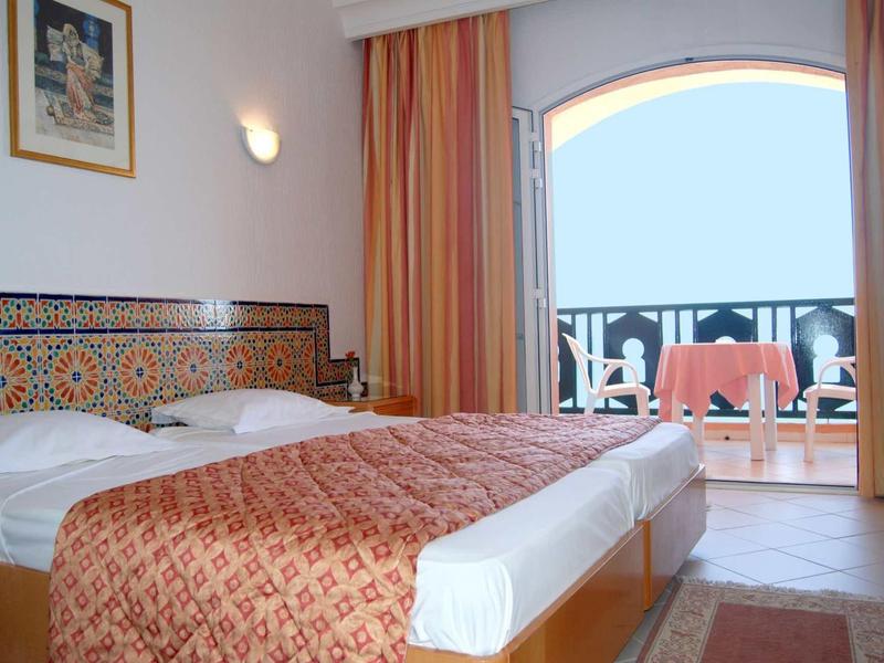 Hell eingerichtetes Schlafzimmer mit Doppelbett, orangen Vorhängen und Balkon mit Meerblick.