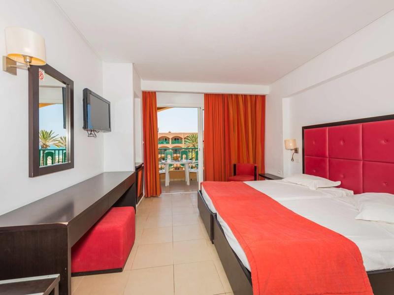 Modernes Hotelzimmer mit Doppelbett, rotem Bettkopfteil und Vorhängen, Schreibtisch und Balkon.