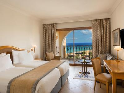 Jasny pokój hotelowy z dwoma łóżkami, balkonem i widokiem na morze przy bezchmurnym niebie.