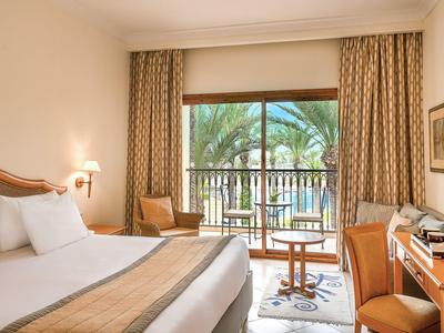 Pokój hotelowy z łóżkiem podwójnym, drewnianymi meblami i widokiem na palmy z balkonu.