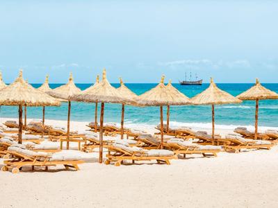 Strand mit weißen Liegen und Strohdächern, blauer Himmel und Meer, Schiff am Horizont.
