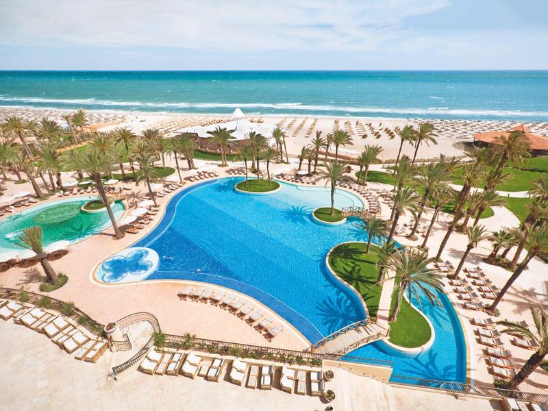 Duży hotelowy basen z palmami i bezpośrednim dostępem do piaszczystej plaży i morza.