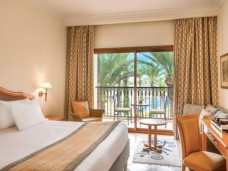 Pokój hotelowy z łóżkiem podwójnym, drewnianymi meblami i widokiem na palmy z balkonu.