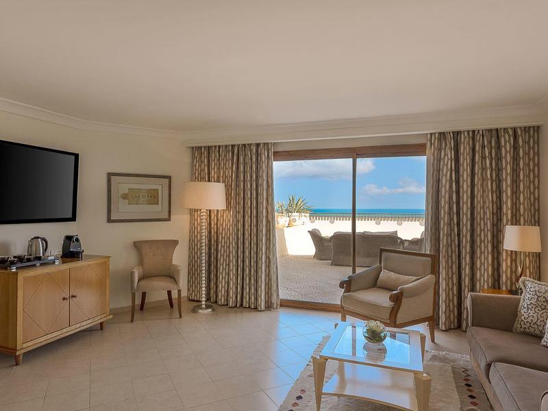 Jasny salon z panoramicznym oknem, beżową sofą, fotelami i telewizorem na ścianie.