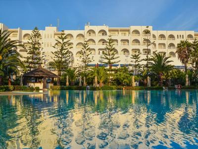 Gran hotel de varios pisos con palmeras y una amplia piscina al frente bajo un cielo despejado.
