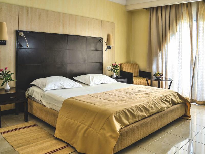 Moderní hotelový pokoj s manželskou postelí, nočními stolky, křesly a velkým oknem se závěsy.