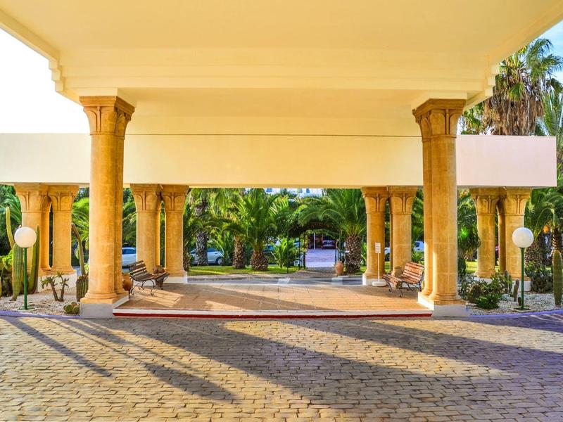 Velký zastřešený vstup s sloupy a zelení za slunečného dne.