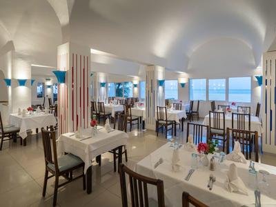 Světle zařízená jídelna s bílými stoly a dřevěnými židlemi, velké okno s výhledem na moře.