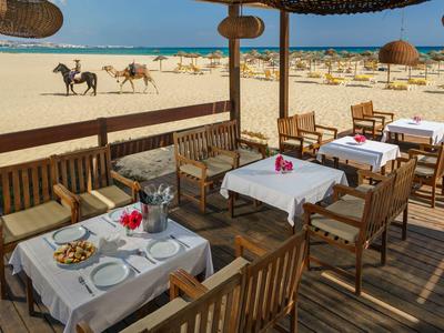 Restaurace s terasou dřevěného nábytku s výhledem na pláž s koňmi v pozadí.