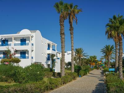 Edificio alberghiero bianco accanto a un sentiero lastricato con palme e piante fiorite sotto un cielo azzurro limpido.