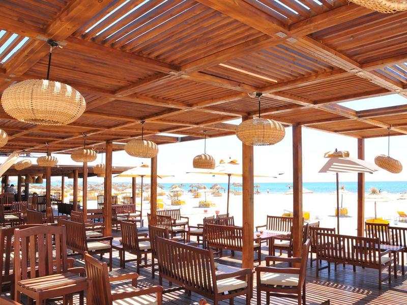 Otevřený plážový bar s dřevěným nábytkem a doškovou střechou, výhled na slunečníky a moře.