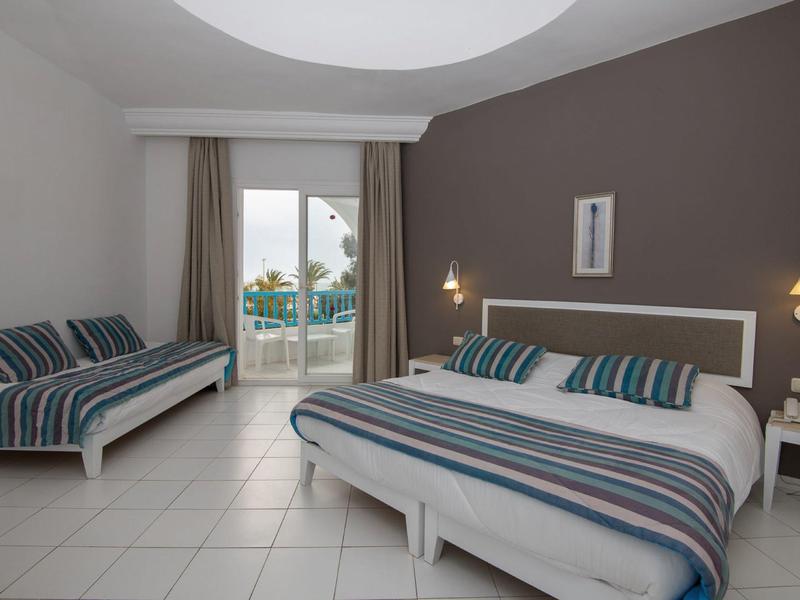 Hotelový pokoj s manželskou postelí, pohovkou, šedou akcentní zdí a výhledem na terasu a moře.