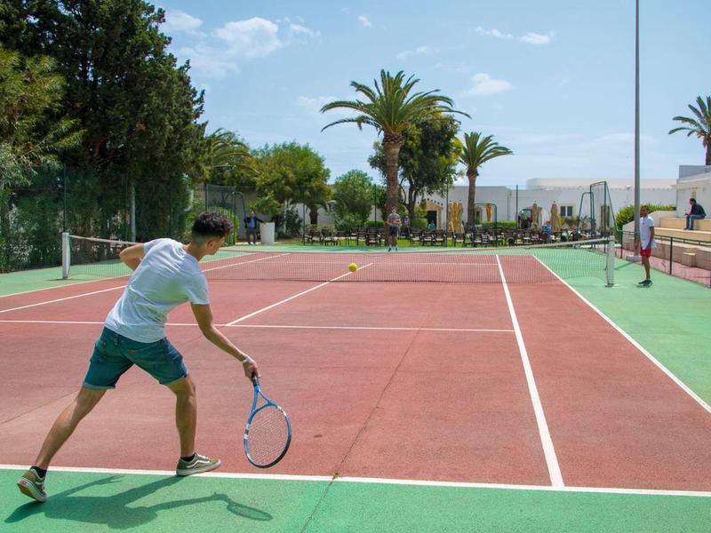 Osoba hrající tenis na venkovním kurtu za slunečného počasí s palmami v pozadí
