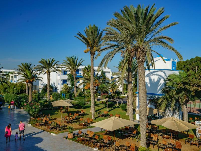 Palmy a posezení před bílou hotelovou budovou pod jasnou modrou oblohou.