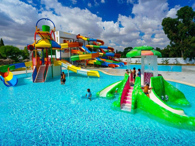 Barevný vodní park s tobogány a několika lidmi v bazénu pod zataženou oblohou.