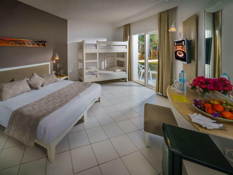 Moderní hotelový pokoj s dvoulůžkem, patrovou postelí, stolem a balkonem s výhledem na zeleň.