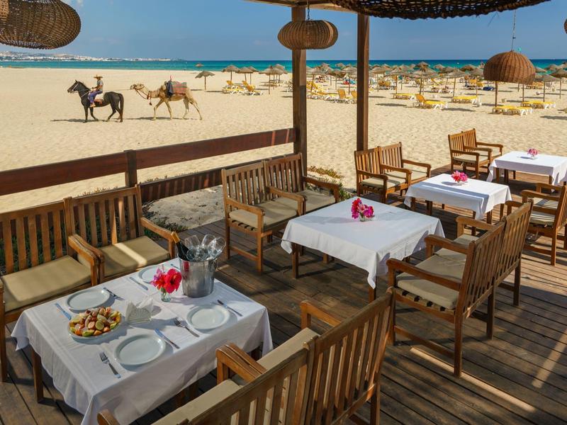 Restaurace s terasou dřevěného nábytku s výhledem na pláž s koňmi v pozadí.