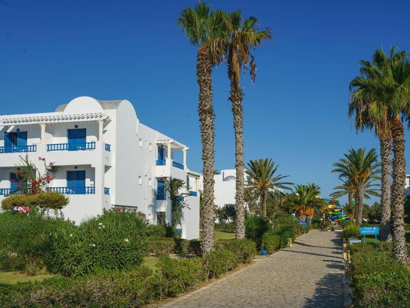 Kamená cesta s palmami a bílými budovami pod jasnou modrou oblohou.