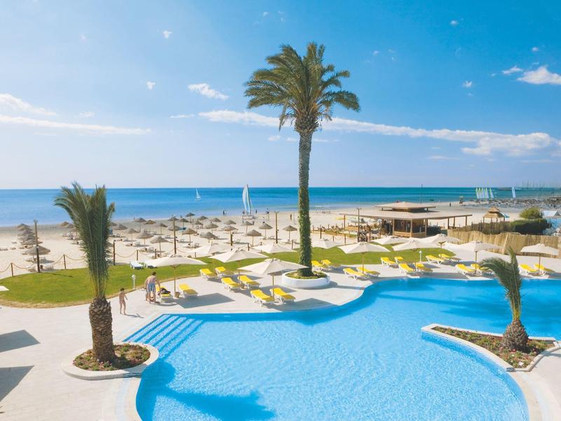 Hotelový bazén s palmami na pláži a slunečníky pod modrou oblohou