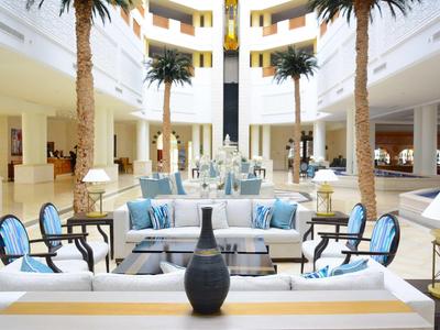 Bright lobby with white sofas, blue cushions, and tall palm trees in a hotel.